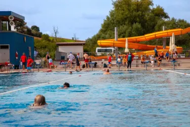 Ein Schwimmbecken mit vielen Menschen, die schwimmen und sich entspannen. Im Hintergrund sind Rutschen und Sonnenschirme zu sehen.