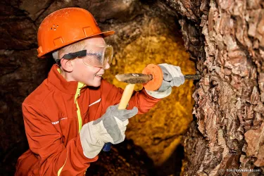 Ein junger Junge arbeitet in einer Mine und nutzt einen Hammer und einen Meißel. Er trägt einen Sicherheitshelm und Schutzbrille.