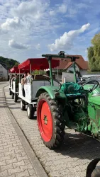 Ein grüner Traktor zieht einen Wagen mit Passagieren. Im Hintergrund sind Häuser und ein blauer Himmel mit Wolken zu sehen.
