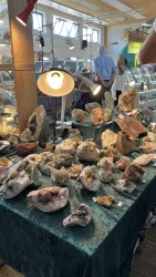 Ein Tisch mit verschiedenen Mineralien und Kristallen, gut beleuchtet mit einer Lampe. Im Hintergrund sind Menschen zu sehen, die die Ausstellung besuchen.