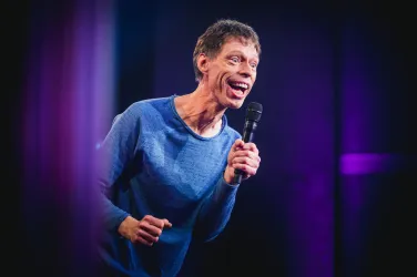 Ein Comedian steht auf der Bühne und spricht ins Mikrofon. Er lächelt und hat eine lebhafte Ausstrahlung.