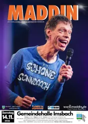 Ein fröhlicher Komiker auf der Bühne, der sein Publikum mit einem Lächeln begeistert. Er trägt ein blaues Shirt mit dem Text „SCHÖNE SONNENDAH“.