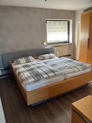 Ein helles Schlafzimmer mit einem großen Bett und karierten Bettdecken. Der Raum hat Holzfußboden und ein Fenster mit Jalousien.