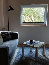 Ein gemütlicher Wohnbereich mit einem grauen Sofa und einem kleinen weißen Tisch. Durch das Fenster scheint Tageslicht und bietet einen Blick ins Grüne.