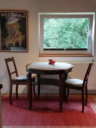 Ein kleiner Tisch mit zwei Stühlen steht vor einem Fenster, durch das viel grünes Licht fällt. Auf dem Tisch liegt eine dekorative Schale mit einem Apfel.