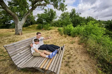Eine gemütliche Holzbank steht in der Natur, umgeben von Grün. Ein Erwachsener und ein Kind sitzen zusammen und schauen in die Ferne.