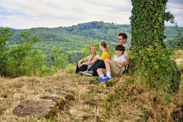 Eine Gruppe von vier Personen sitzt entspannt auf dem Boden in der Natur. Im Hintergrund sind sanfte Hügel und Bäume sichtbar.