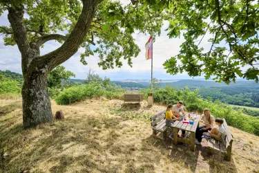 Eine Gruppe von Menschen sitzt an einem Holztisch unter einem Baum. Im Hintergrund ist eine schöne Landschaft mit Hügeln und einem Fahnenmast zu sehen.