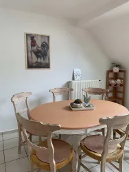 Ein einladender Essbereich mit einem runden Tisch und Holzhockern. An der Wand hängt ein Bild eines Huhns, während ein Regal mit Geschirr im Hintergrund steht.
