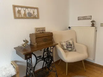 Ein gemütlicher Raum mit einem bequemen Sessel und einer alten Nähmaschine. An der Wand hängen dekorative Gegenstände und ein Schild.