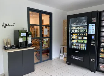 Ein moderner Aufenthaltsraum mit einem Verkaufsautomaten für Getränke und Snacks. Auf der linken Seite steht eine Kaffeemaschine und eine offene Tür führt in einen weiteren Raum.
