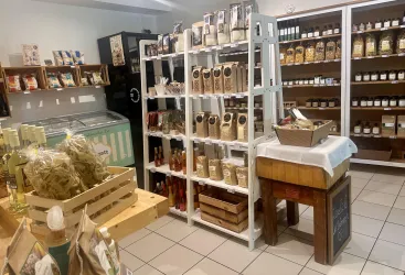 Ein gemütlicher Laden mit Regalen voller Lebensmittel und Produkten. Im Vordergrund steht ein Holztisch mit Waren und einer Kiste.