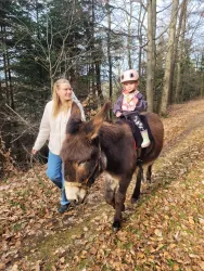 Ein Mädchen sitzt auf einem Esel und trägt einen Helm. Eine begleitende Frau führt den Esel auf einem Waldweg.