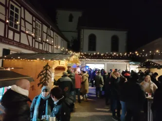 Ein festlicher Marktplatz mit stimmungsvoller Beleuchtung und vielen Menschen. Stände bieten verschiedene Waren an und die Atmosphäre ist gemütlich.