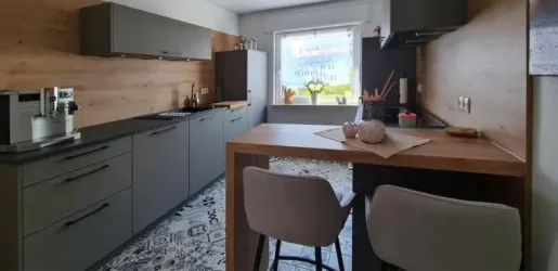 Eine moderne Küche mit grauen Schränken und Holzverkleidungen. Ein Esstisch mit Stühlen steht im Vordergrund, und ein Fenster bietet Tageslicht.