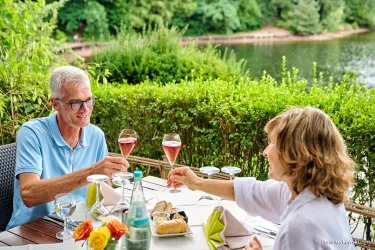 Ein Paar stößt mit Gläsern an und genießt eine Mahlzeit im Freien. Im Hintergrund ist ein ruhiger See und grüne Pflanzen zu sehen.
