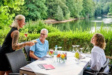 Eine freundliche Kellnerin serviert Getränke an einem Tisch mit Blick auf einen schönen See. Die Gäste wirken entspannt und genießen die Atmosphäre in der Natur.