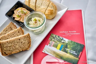 Ein Teller mit frischem Brot, einem Dip und verschiedenen Beilagen. Darauf liegt eine Menükarte des Restaurants "Seehaus Forelle Haeckenhaus".