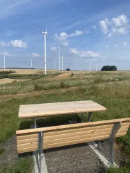 Ein ruhiger Aussichtspunkt mit einem Holzstuhl und Tisch. Im Hintergrund sind Windräder und eine hügelige Landschaft zu sehen.