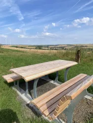 Ein Holzpicknicktisch steht in einer grünen Wiese mit Blick auf Felder und einen blauen Himmel. Sanfte Hügel und einige Windräder sind im Hintergrund sichtbar.