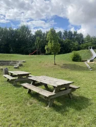 Ein Spielplatz mit einer Rutsche und einem Klettergerüst im Hintergrund. Im Vordergrund stehen Holzpicknicktische auf einer grünen Wiese.