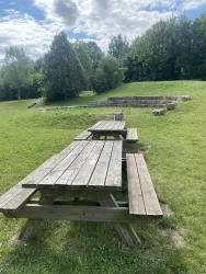 Ein Holzpicknicktisch steht in einer grünen Wiese. Im Hintergrund sind Bäume und ein kleiner Hügel zu sehen.