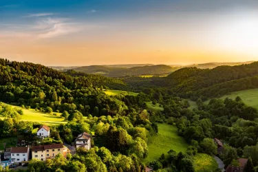 Eine schöne Landschaft mit sanften Hügeln und üppigem Grün. Im Hintergrund sieht man einen Sonnenuntergang über den Bergen.