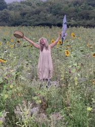 Eine Frau steht begeistert in einem Sonnenblumenfeld. Sie hält einen Hut und einen Schirm in den Händen.