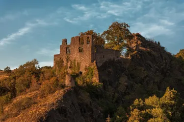 Eine alte Burgruine auf einem Hügel, umgeben von grünen Bäumen. Der Himmel ist klar mit sanften Wolken und das Licht ist warm.