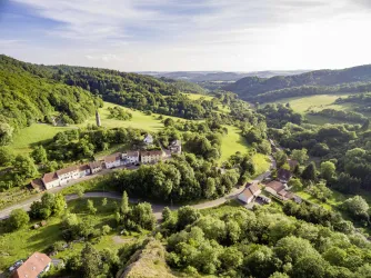 Eine malerische Landschaft mit sanften Hügeln und üppigen Wäldern. Im Vordergrund sind einige Häuser und eine kurvenreiche Straße zu sehen.