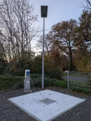 Eine öffentliche Ladestation für Elektrofahrzeuge in einem parkähnlichen Bereich. Zu sehen sind Bäume im Hintergrund und ein grauer Boden mit einem Gitter.