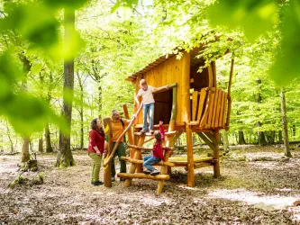 Eine Familie spielt um ein Baumhaus im Wald. Die Umgebung ist grün und sonnig, ideal für Aktivitäten im Freien.