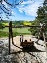Eine Schaukel steht auf einem Anhöhen mit Blick auf eine grüne Landschaft. Im Hintergrund blühen gelbe Rapsfelder unter einem blauen Himmel.