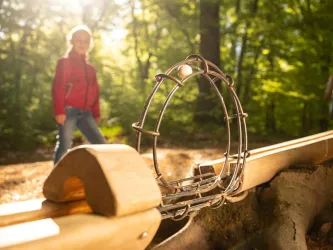 Ein Spielgerät im Wald aus Holz und Metall. Im Hintergrund ist ein Kind in einem roten Kleidungsstück zu sehen.