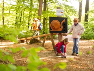 Ein Spielplatz im Wald mit einem lustigen Schnecken-Schild. Ein Mädchen bindet ihre Schuhe, während ein Erwachsener im Hintergrund spaziert.