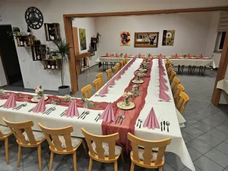 Ein festlich gedeckter Tisch in einem Restaurant mit weißen Tischdecken und rosa Servietten. Die Stühle sind aus Holz und es gibt dekorative Elemente auf dem Tisch.