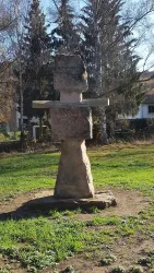Eine große Steinskulptur steht auf einer Wiese. Im Hintergrund sind Bäume und ein Gebäude zu sehen.