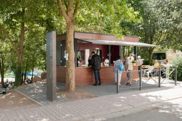 Ein moderner Imbissstand unter Bäumen mit Gästen, die essen und sich unterhalten. Die Umgebung ist hell und einladend.