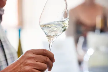 Ein Glas Weißwein wird in der Hand gehalten. Im Hintergrund sind unscharfe Personen und eine entspannte Atmosphäre zu sehen.