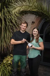 Ein Mann und eine Frau stehen vor einer Pflanze und halten eine Flasche. Beide lächeln und wirken freundlich.