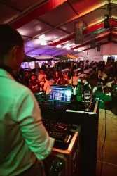 Ein DJ steht an einem Mischpult und spielt Musik in einem Zelt voller Menschen. Die Atmosphäre ist lebhaft und festlich, mit bunten Lichtern und Getränken auf den Tischen.