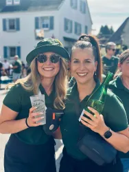 Zwei Frauen lächeln fröhlich in einer belebten Umgebung. Beide tragen grüne Hemden und halten Getränke in ihren Händen.