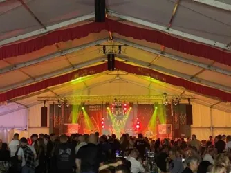 Ein überfülltes Zelt mit Musikern auf der Bühne und bunten Lichtern. Die Atmosphäre ist festlich und lebhaft, mit vielen Menschen, die feiern.