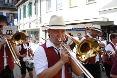 Eine Blaskapelle marschiert durch die Straße. Die Musiker tragen traditionelle Kleidung und spielen verschiedene Instrumente.