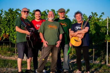 Eine Gruppe von fünf Musikern steht in einem Weinberg. Sie tragen T-Shirts mit verschiedenen Farben und halten Musikinstrumente in den Händen.