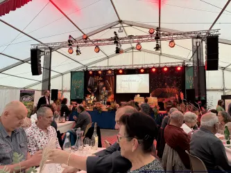 Eine festliche Veranstaltung in einem Zelt mit vielen Gästen an Tischen. Eine Bühne ist im Hintergrund mit Beleuchtung und einer Präsentation zu sehen.