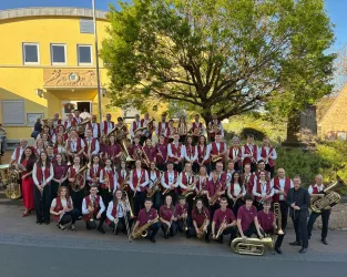 Eine große Blaskapelle steht vor einem gelben Gebäude unter einem großen Baum. Die Musiker tragen einheitliche Kleidung in Rot und Weiß und präsentieren verschiedene Instrumente.