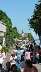 Eine belebte Straße mit vielen Menschen und Ständen. Im Hintergrund ist ein Kirchturm sichtbar.