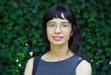 Eine Frau mit dunklen, lockigen Haaren und einer Brille steht vor einem grünen Hintergrund. Sie trägt ein schlichtes, ärmelloses Kleid und lächelt leicht.