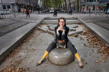 Eine Person sitzt auf einem runden Stein in der Stadtmitte und lächelt. Um sie herum sind Fußgänger und Bäume zu sehen.